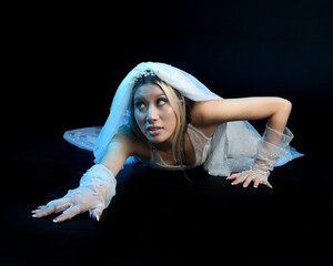Wall Mural - Full length portrait of beautiful model wearing white wedding gown and ghostly flowing veil like a shroud. Moody cinematic lighting, sitting pose, isolated on a dark studio background.