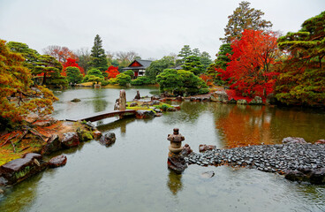 Wall Mural - Fall scenery of fiery maple trees by a lake & a stone bridge over the pond on a drizzling day in the beautiful Japanese garden of Katsura Rikyu Imperial Villa, a detached Royal Palace in Kyoto, Japan