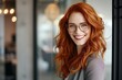 © Rudsaphon - Bright and inviting portrait of a red-haired woman with glasses, smiling confidently in a professional office environment.