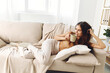 © SHOTPRIME STUDIO - Cozy mug of happiness: A young, smiling woman sitting alone on a sofa in a warm and modern living room, holding a cup of coffee with a blanket draped over her relaxed body.