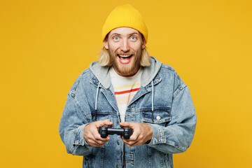 Young gambling happy man he wearing denim shirt hoody beanie hat casual clothes hold in hand play pc game with joystick console isolated on plain yellow background studio portrait. Lifestyle concept.