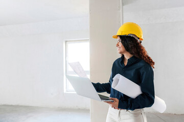 Smiling architect holding laptop and examining site with blueprints