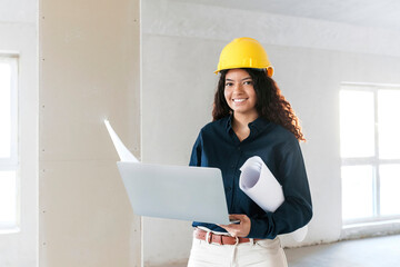 Smiling young architect holding laptop and blueprints at site