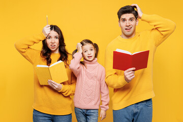 Wall Mural - Young sad shocked parents mom dad with child kid girl 7-8 years old wear pink sweater casual clothes read book help study with difficult task isolated on plain yellow background. Family day concept.