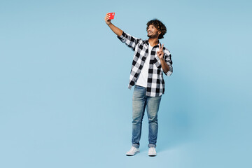 Wall Mural - Full body young Indian man wear shirt white t-shirt casual clothes doing selfie shot on mobile cell phone post photo on social network show v-sign isolated on plain blue background. Lifestyle concept