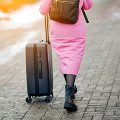  Woman in pink coat and boots walking with suitcase on wheels. Female legs and luggage on the street, travel concept. Tourist with suitcase walk along snowy street in winter, selective focus