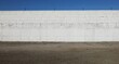 Long white concrete block wall with barbed wire on top. Asphalt street in front, blue sky above. Background for copy space. 