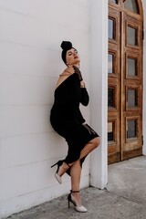 Wall Mural - Stylish woman in the city. Fashion photo of a beautiful model in an elegant black dress posing against the backdrop of a building on a city street