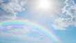 © Nikki Zalewski - Soft beautiful rainbow sky spiritual background -  blue sky with pretty clouds, a bright sun shining center top and a large rainbow arcing from left to right with copy space