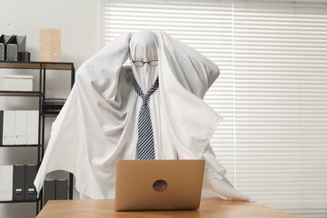 Overworked ghost. Man covered with white sheet using laptop at wooden table in office