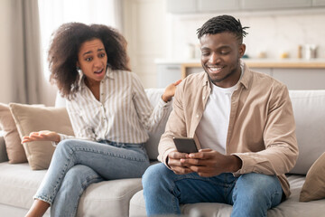 Wall Mural - Discontented Black Wife Looking At Husband Browsing On Phone Indoor