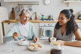 Fototapeta  - Indoor portrait of two people of diverse age and ethnicity sitting at kitchen table, african american young woman drinking tea with elderly caucasian female treating her with sweets