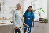 Fototapeta  - Side view of black african american nurse volunteer teaching elderly woman with gray hair in cardigan to use walker, supporting her and giving advice and instructions, standing in her kitchen