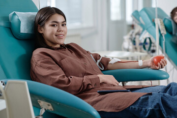 Wall Mural - Smiling young woman sitting in comfortable chair in medical center and donating blood