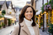 Portrait einer lächelnden, braunhaarigen Frau mit beigem Mantel und hellem Pullover in einer kleinen Einkaufsstraße