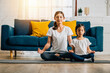© sorapop - A beautiful young woman and her charming daughter share smiles during their family yoga session at home emphasizing mindfulness and meditation in lotus position creating happiness and togetherness.