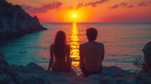  A Man And A Woman Sitting On A Rock Looking Out At The Ocean As The Sun Sets Over The Water.