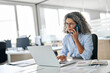 © insta_photos - Busy mature middle aged business woman, senior professional lady executive manager talking on the phone making business call on cellphone sitting at work desk in office using laptop computer.