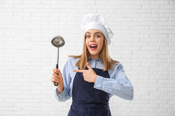 Poster - Young female chef pointing at ladle on white brick wall background