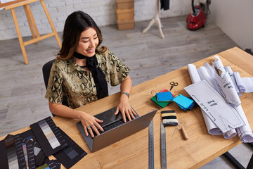 Wall Mural - high angle view of cheerful asian stylist looking at laptop near color samples on desk in atelier