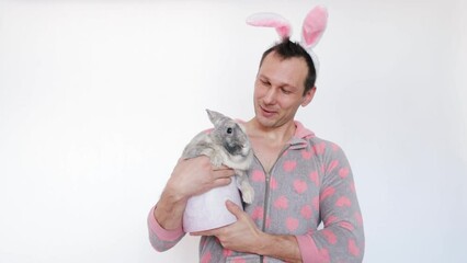 Wall Mural - Portrait of the smiling handsome man holding the fluffy easter rabbit, wearing bunny ears. Happy male is getting ready for the spring holiday looking at camera. Preparation for easter.