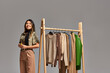 © LIGHTFIELD STUDIOS - pleased asian stylist with measuring tape looking away near rack with bespoke clothes on grey