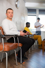 Wall Mural - Adult man in casual clothes waiting for his turn while sitting on chair with phones in his hand