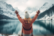 Travel Background Concept. The Back View Of The Woman With Hands Up, Looking And The Beautiful Landscape With The Lake Or The River, Mountains Covered In Snow, Trees In Winter