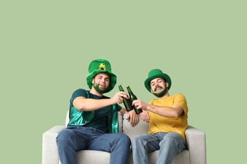 Wall Mural - Young men in leprechaun hats with green beards holding bottles of beer and sitting on sofa against green background. St. Patrick's Day celebration