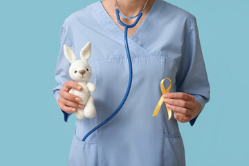 Wall Mural - Doctor holding golden ribbon and toy bunny on blue background. Childhood cancer awareness concept