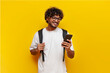 © Богдан Маліцький - young indian guy student in a white t-shirt with glasses and with a backpack uses a smartphone and selects online on a yellow isolated background, a curly-haired man types a message on a mobile