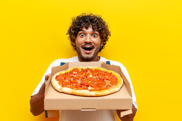 Wall Mural - young shocked indian man holding a box of delicious pizza and wondering on yellow isolated background, curly guy student showing and advertising fast food in amazement with open mouth