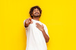 young indian man in a white t-shirt mocks and jokes and points his finger forward on a yellow isolated background, curly-haired guy laughs and ridicules and mocks