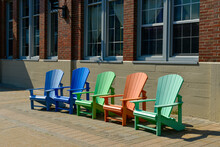 A Row Of Colorful Recycled Wood Adirondack Chairs On A Sidewalk Next To A Large Red Brick Building With Windows. The Resting Area Has Blue, Green, Orange And Teal Colored Plastic Stackable Deck Chairs