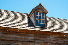 An Old Dormer Window With Multiple Panes Of Glass, White Trim And A Peaked Overhang. The Roof Is Made Of Cedar Shakes. The Red Wall Of The Abandoned House Has Peeling Paint And Is Worn And Weathered. 