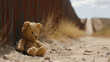 A lone teddy bear sits against a border fence, a silent symbol of childhood disrupted by migration.