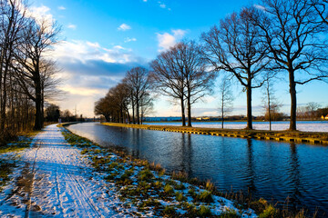 Wall Mural - This captivating image showcases a snow covered pathway running alongside a tranquil canal, flanked by majestic, leafless trees against a bright winter sky. The setting sun casts a soft light