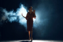 From Shadows, Voice Takes Flight. Woman Singing Into Microphone Surrounded Dramatic Smoke And Backlighting On Stage Against Black Background. Performance. Concept Of Hobby, Festival, Concert, Disco.
