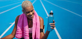Fototapeta Koty - Smiling female athlete drinking energy drink from bottle after running sport exercise, wears earphones, smartphone armband and towel. African American woman sitting on Olympic track with blue lanes
