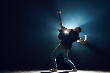 Rear view portrait of street style musician strumming chords on black guitar with dramatic on stage against black background with backlights. Concept of Rock-n-roll, music and dance, culture.