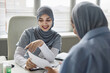 © Seventyfour - Cheerful young Islamic woman physician in gray headscarf talking to unrecognizable female patient looking at medical document in hand and sitting at table in clinic