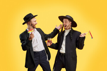 Two Jewish man, friends happy to meet at Purim holiday celebration. Male model posing with grogger and alcohol drinks against yellow background. Festival, Judaism, religion concept. Ad