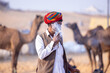 Portrait of an old male from rajasthan in traditional white dress and colourful turban with camel smoking at desert fair ground during pushkar fair.	
