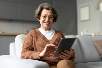 Delighted senior woman enjoy using digital tablet on sofa, radiating happiness and contentment, showing harmonious blend of technology and comfort