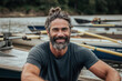A casual, bearded man with a top knot hairstyle smiles warmly by a river, with rowboats in the background, suggesting an active, outdoor lifestyle