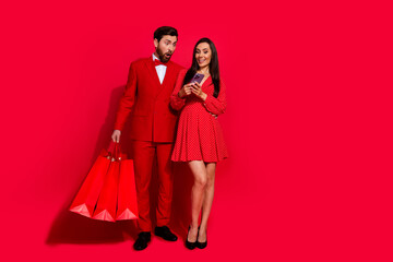 Wall Mural - Full body photo of two elegant beautiful impressed people hold shop bags use smart phone empty space isolated on red color background