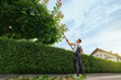 © serhiibobyk - Strong caucasian man in uniform and gloves using petrol trimming machine for cutting overgrown trees outdoors. Gardener landscaping during summer time.
