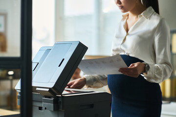 Wall Mural - Cropped shot of young pregnant businesswoman in quiet luxury attire putting paper on copier screen of xerox machine in office