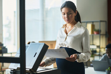Poster - Young pregnant Asian businesswoman in smart casualwear looking at financial document while standing by xerox machine in office