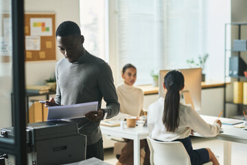 Poster - Young African American office manager in casualwear looking at financial document while standing by xerox machine against colleagues